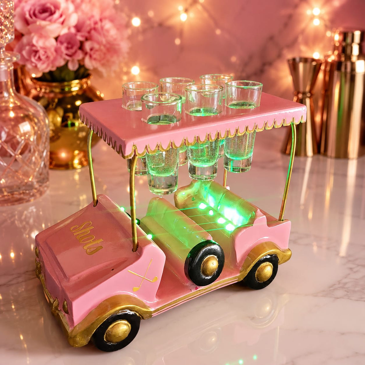 A pink golf cart-shaped bar table with gold trim and multiple clear shot glasses on top, lit with green LED lights, set on a marble surface with warm decorative background elements.