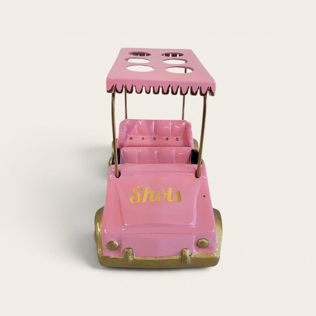 A pink novelty golf cart toy or decor piece with gold-colored wheels and trim, featuring the word 'Shots' in gold script on the front. The canopy has cutouts, and the cart is photographed on a clean, light beige background.