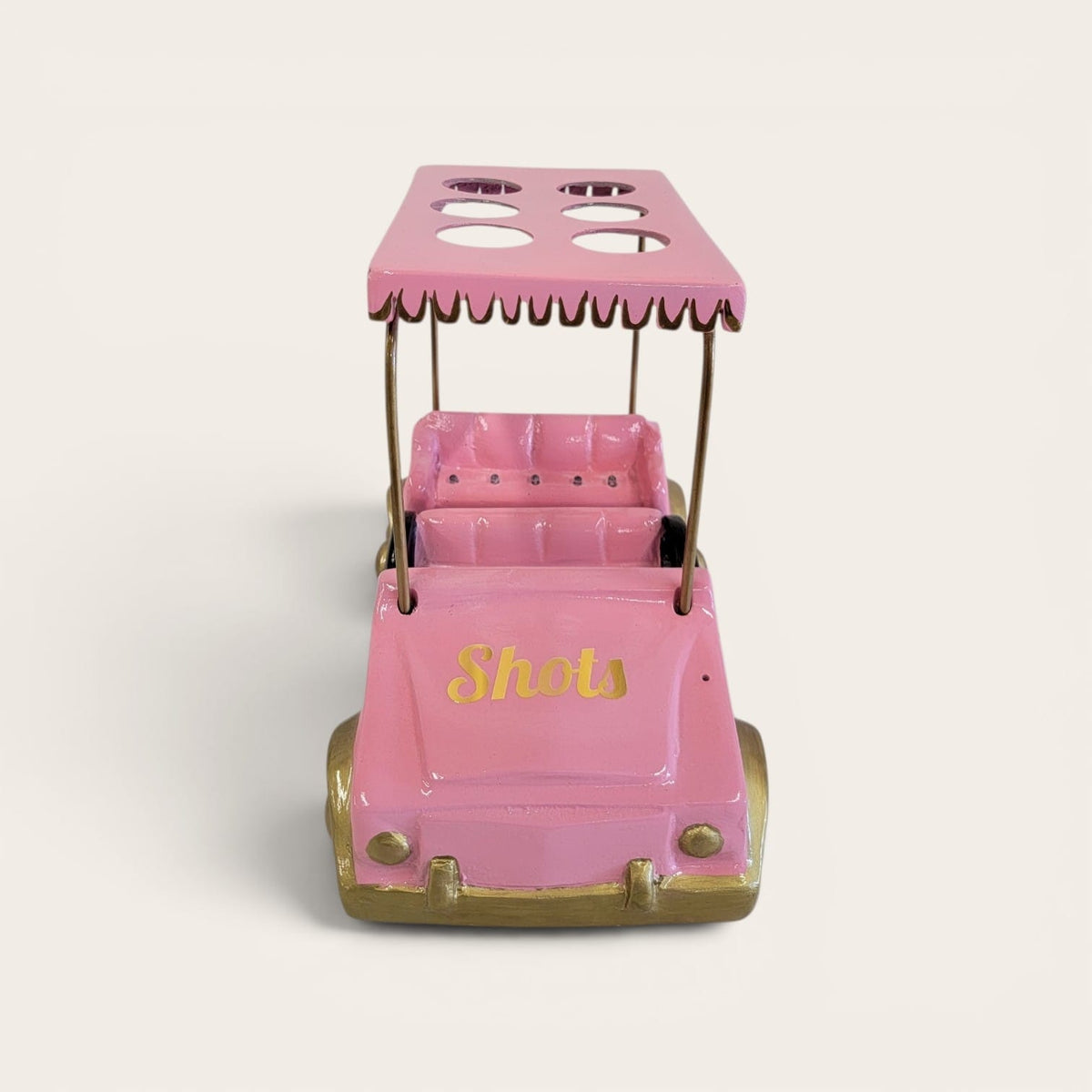 A pink novelty golf cart toy or decor piece with gold-colored wheels and trim, featuring the word 'Shots' in gold script on the front. The canopy has cutouts, and the cart is photographed on a clean, light beige background.
