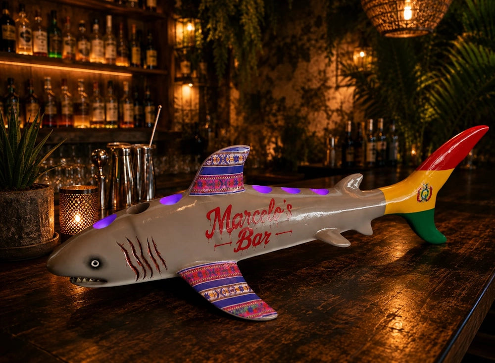 A decorative shark bar sign with 'Marcela's Bar' written on its side, displayed on a dark wooden bar counter in a warmly lit bar setting.