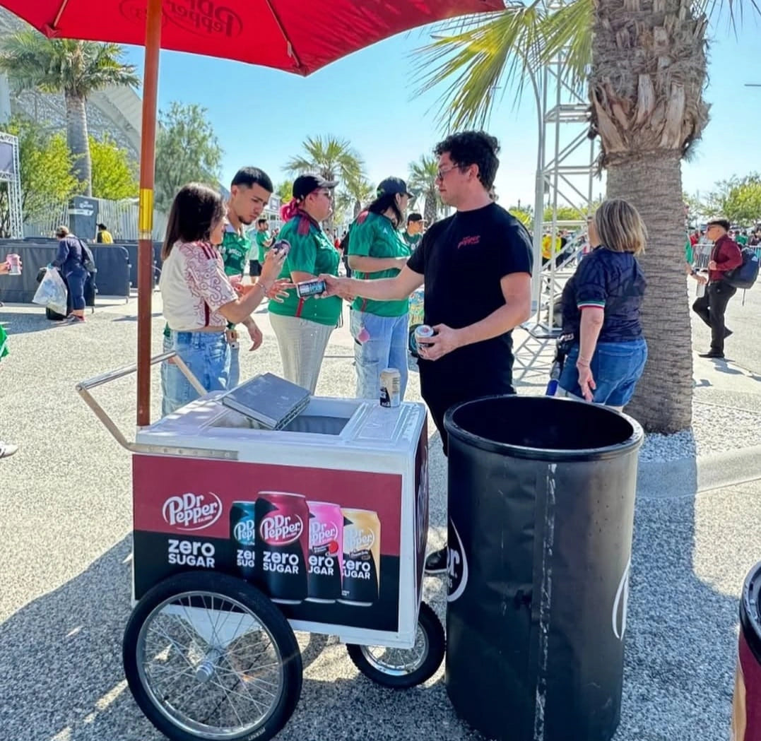 A Dr Pepper zero sugar beverage cart at an outdoor event, with people standing around and a large black trash can in the foreground under a red umbrella.