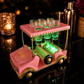 A pink golf cart-shaped shot glass holder with gold trim and six clear shot glasses on top, illuminated with green LED lights on a dark reflective surface.