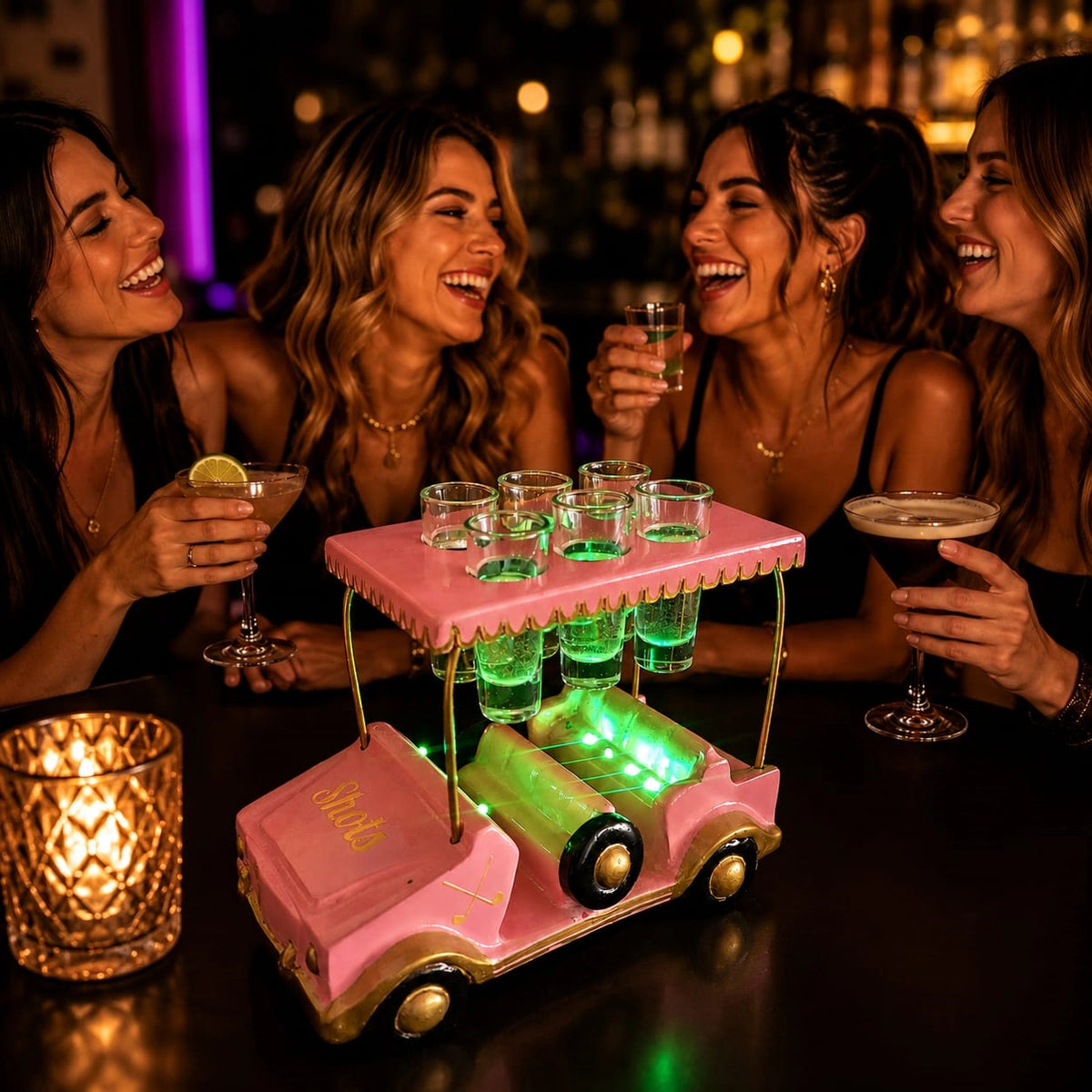 A pink and gold car-shaped bar cart filled with shot glasses and glowing green LED lights, set on a dark bar table with four smiling women holding cocktails in the background.