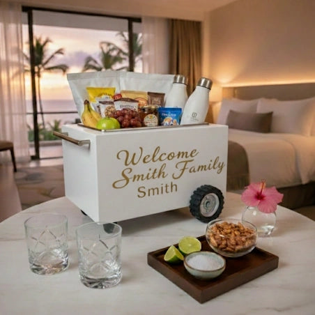 A customized white gift cart filled with snacks, drinks, and fruit, parked on a table in a hotel room. The cart is personalized with the text 'Welcome Smith Family Smith' in gold script.