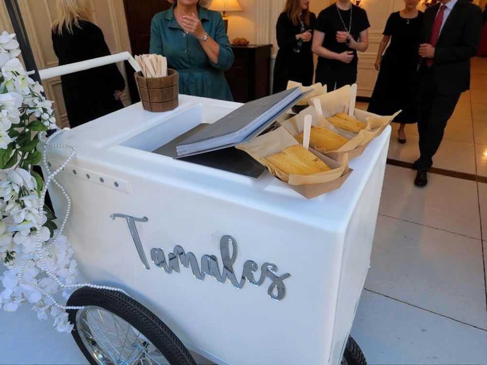 A white wheeled food cart with the word 'Tamales' in script on the side, set in an indoor event space. Papers and small food items are arranged on top, with guests standing in the background.