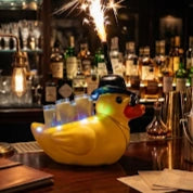 A yellow rubber duck novelty candle or light on a bar counter, wearing a black top hat and smoking a sparkler, with a blurred bar background.